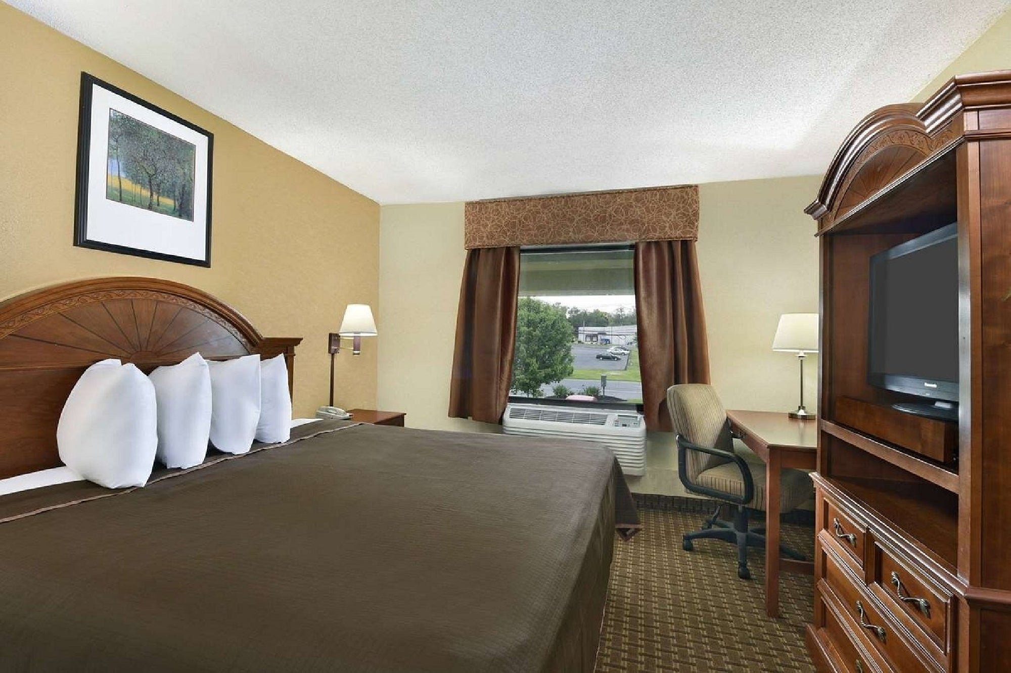 Hotel room with a large bed, four pillows, wooden furniture, a desk with a chair, TV, and a window with a brown curtain showing a view of a parking lot and greenery outside.