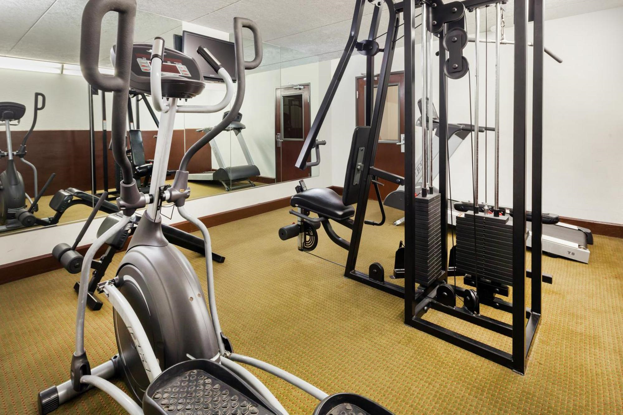 A small gym room with an elliptical machine, a weight machine, a treadmill, a mirror along one wall, and a closed door in the background.