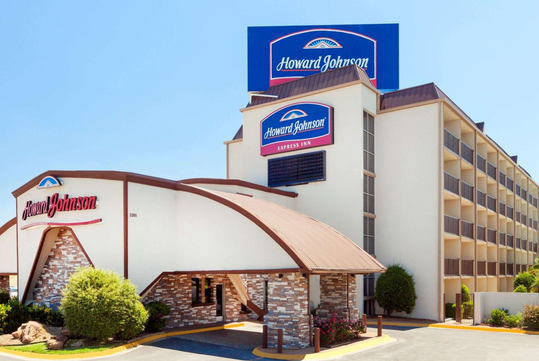 Exterior of a Howard Johnson hotel with a prominent blue sign, multiple floors, balconies, and landscaped shrubs in front.
