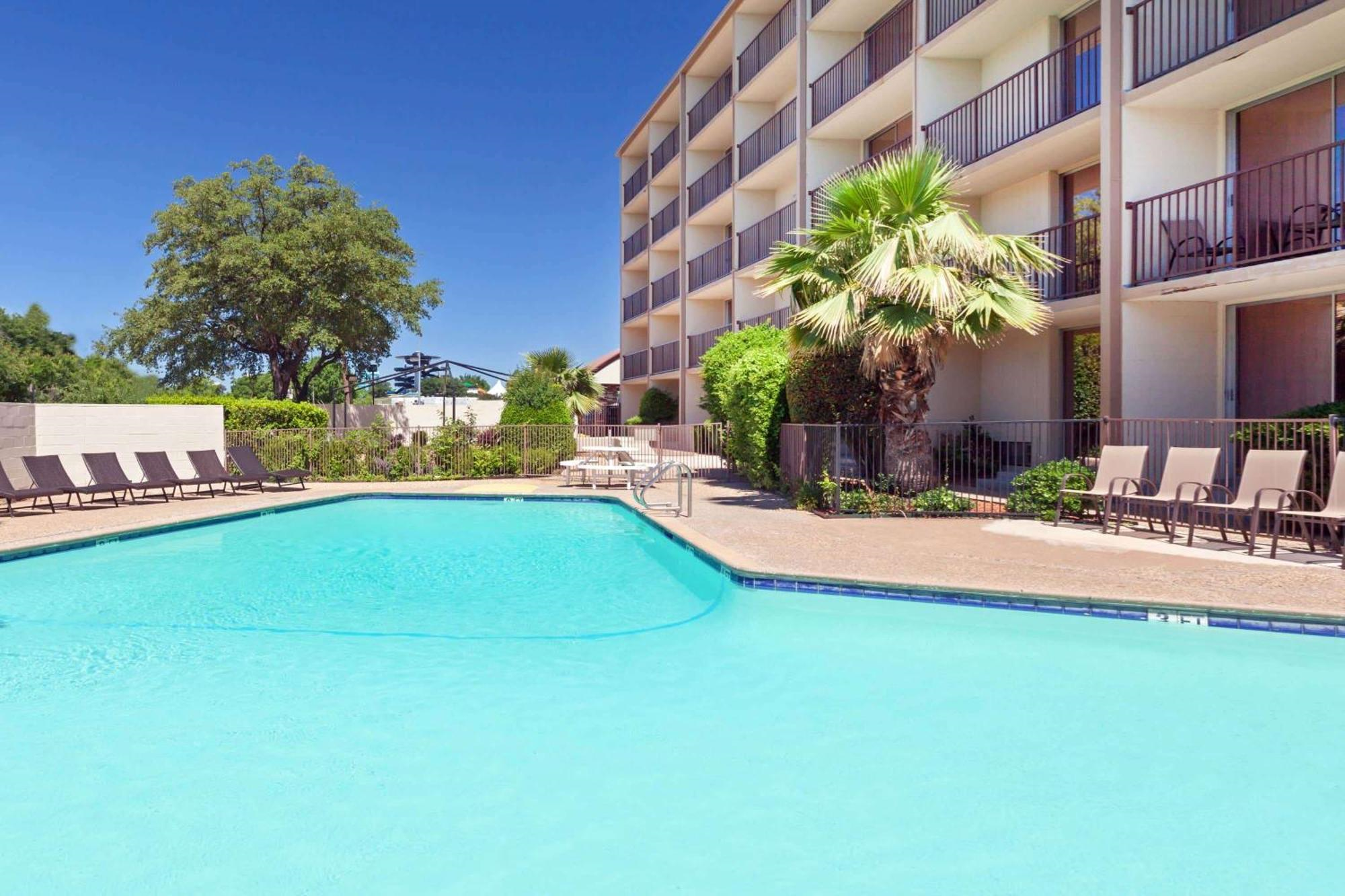Outdoor swimming pool next to a multi-story hotel building with lounge chairs, palm trees, and landscaping under a clear blue sky.