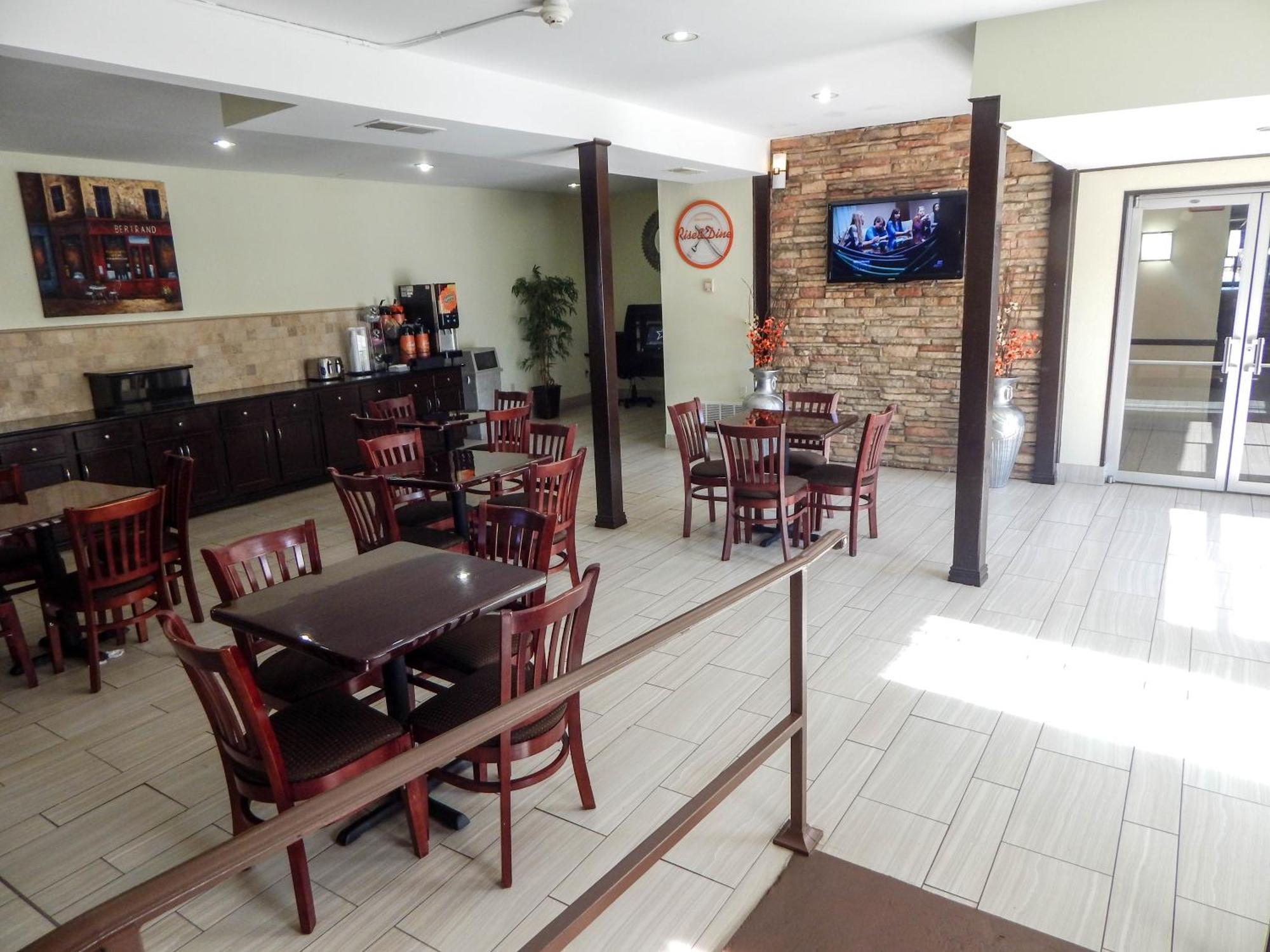 Hotel breakfast area with several dark wood tables and chairs, a TV on a stone wall, a coffee station, and large windows letting in natural light.