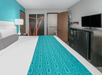 A hotel room with a white king bed with a blue runner, a TV on a stand with a mini fridge under it and a bathroom on the far wall.