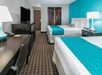 A hotel room with two white and blue queen beds, a desk, a side table with lamp, and a TV on a stand with a mini fridge under it.