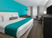 A hotel room with a white and blue king bed, a desk, a love seat, and a TV on a stand with a mini fridge under it.