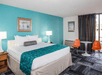 Hotel room with a king-size bed, teal accent wall, and geometric-patterned bed runner. Two orange chairs and a round table are in the corner, next to a window with striped curtains.