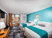 Hotel room with two double beds featuring teal accents, a flat-screen TV, a wooden dresser, and a small seating area by a window with orange chairs. A poolside view is visible outside.