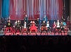 A group of performers on stage, dressed in colorful and coordinated costumes, sing and dance in front of a red and white backdrop with dramatic lighting.