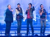 Four men sing on a brightly lit stage, each holding a microphone. Stage lights and musical instruments are visible in the background.