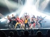 A group of performers pose energetically on stage in colorful outfits, with bright stage lights shining behind them and an audience watching.