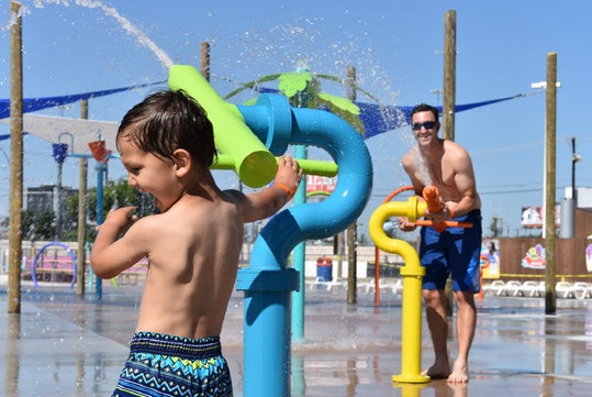 A child and an adult play with colorful water cannons at an outdoor splash pad on a sunny day.