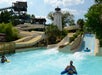 People on inflatable rafts enjoying water slides at a water park with a lighthouse structure and surrounding greenery.