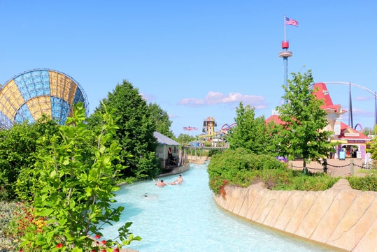 Amusement park scene with water ride in foreground, two people in a raft, roller coaster, tall tower, and American flag visible in the background under a clear blue sky.