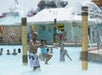 A man lifts a smiling child in a colorful swimsuit in a water park pool, with fountains and other visitors in the background.