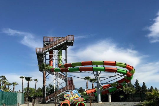 Tall water slide with a twisting, colorful tube structure and stairs leading up, set against a clear blue sky with palm trees nearby.