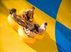 Four people ride together on a large yellow inflatable down a water slide, smiling as they hold onto handles. The slide features bright yellow and blue sections.