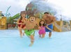 A family of four walks through shallow water at a water park. A boy and a girl run ahead, splashing, while the adults follow, smiling. A large water slide is in the background.