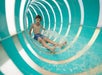 Boy in blue swim trunks sliding down an enclosed, turquoise and white water slide, surrounded by splashing water.