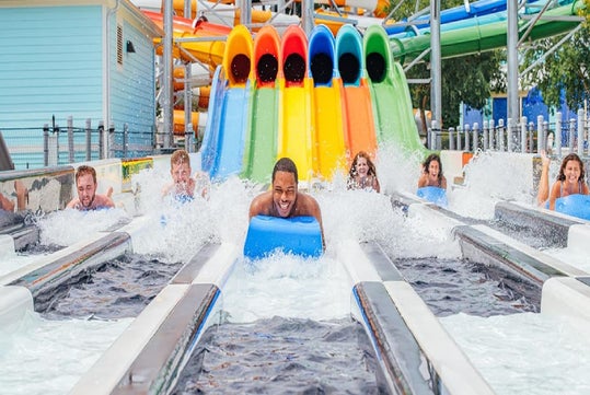 A group of people race down parallel water slides on mats, making splashes as they reach the bottom at an outdoor water park.