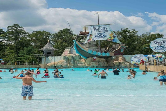 People enjoy a wave pool at a water park with a pirate ship structure. Banners for Welch's Fruit Snacks are visible. Trees and a cloudy sky are in the background.