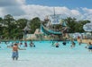 People enjoy a wave pool at a water park with a pirate ship structure. Banners for Welch's Fruit Snacks are visible. Trees and a cloudy sky are in the background.