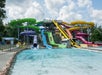 Multiple colorful water slides and chutes empty into a large shallow pool at a water park, with a cloudy blue sky above.
