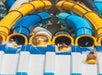 Four people slide down colorful water slides, each in a tube. The slides are yellow, blue, and white, creating a vibrant atmosphere at the water park.