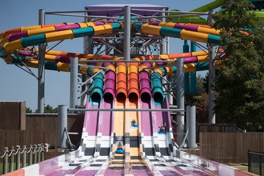 Colorful multi-lane water slide with people sliding down in a water park; vibrant tubes arch overhead while trees and a fence are in the background.