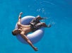 A person relaxes on a white inflatable ring in a pool, wearing floral swim shorts, with arms outstretched and eyes closed, under bright sunlight.