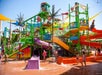 Children play at a colorful water park with various slides and climbing structures. The ground is wet, and there are vibrant decorations, including palm tree motifs and a toucan statue.