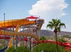 People stand at the top of a yellow and orange waterslide under a red canopy. The slide curves down with mountains and a palm tree in the background.