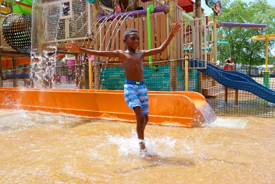 A child in blue swim trunks joyfully runs through a water spray at a colorful playground with slides and climbing structures in the background.