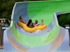 Four people ride a large yellow inflatable raft down a green and blue water slide, surrounded by trees and a parked vehicle in the background.