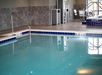 Indoor swimming pool with clear water, metal handrails, and depth markers. Tables and chairs are visible in the background.