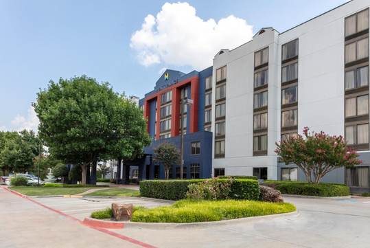 A four-story hotel building with white and dark blue exterior walls, large windows, trees, and landscaping in front, under a partly cloudy sky.