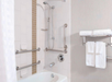 Accessible bathroom with white tiles, a bathtub, grab bars, a handheld showerhead, a towel rack with folded towels, and built-in soap holders.