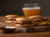 Three pulled pork sliders with coleslaw on a wooden plate with a glass of beer behind them.