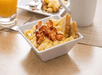 A bowl of scrambled eggs with bacon and toast on top with other breakfast food in the background.