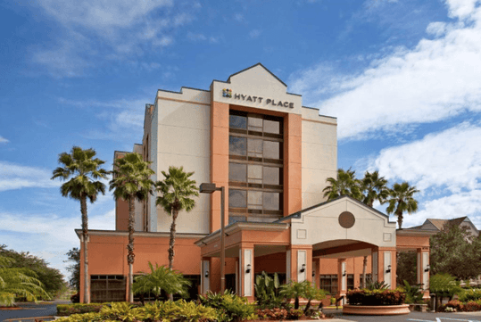 The front exterior of the Hyatt Place Orlando with several palm trees in front of it and a blue cloudy sky overhead.