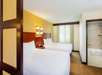Hotel room with two double beds, wooden headboards, and a nightstand with a phone. The room features a window with curtains and an en-suite bathroom with a tub and beige tiles.