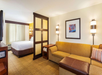 Hotel room interior with a large bed, yellow sofa, wall art, and two partition walls with a window view. Room includes carpet flooring and modern lighting fixtures.