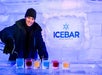 A person pours drinks into ice glasses at ICEBAR Orlando, surrounded by ice walls and blue lighting.