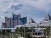 A modern cityscape with high-rise buildings and a unique roofline structure, surrounded by palm trees under a partly cloudy sky.