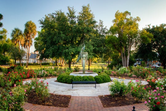 A circular garden features a central sculpture, surrounded by roses and greenery, with trees in the background and a brick walkway in the foreground.