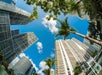 Fisheye view of tall skyscrapers and palm trees against a bright blue sky with scattered clouds.