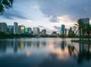 City skyline with tall buildings reflecting on a calm lake at sunset, surrounded by trees and a partly cloudy sky.