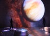 Two people stand in a dark room with a large projection of a planet and star-filled galaxy displayed on the walls and floor.