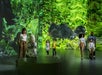 Several people, including adults and children, are in an exhibit space with a large projected image of a gorilla in a lush forest on the wall.