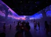 People stand and walk inside a dark room with walls and ceiling displaying a colorful digital landscape of trees and a sunset.