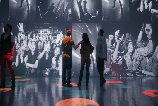 Three people stand in a room with large black-and-white concert photos projected on the walls, depicting energetic crowds and performers.
