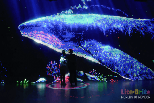 Two people stand in front of a large, colorful digital display showing a pixelated whale underwater with coral, at an exhibit labeled "LiteBrite WORLDS OF WONDER.